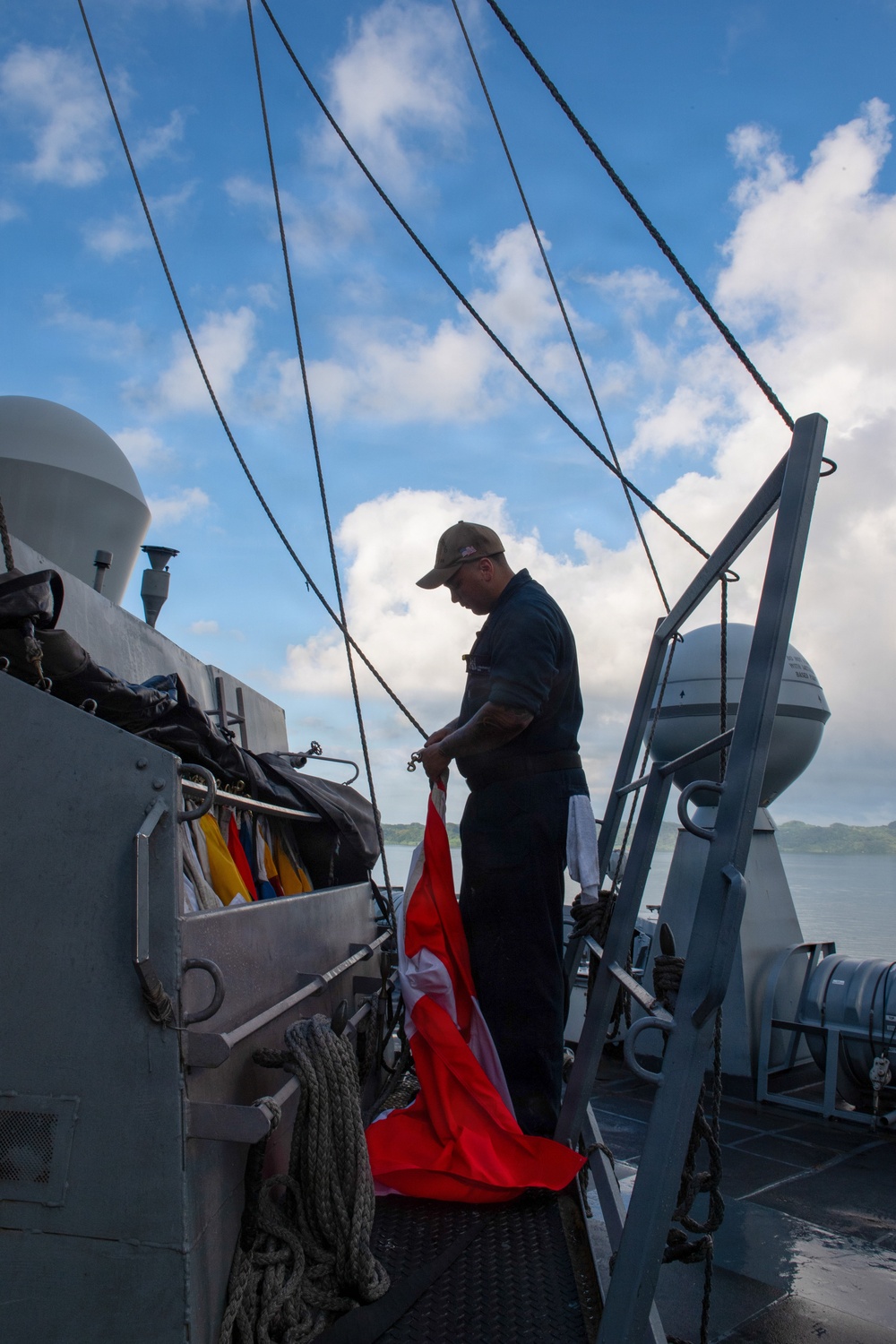 A naval sailor dressed in dark blue uniform and a cap is handling a large red and white flag on the deck of a military vessel. The ship appears to be underway or anchored near a coastline with green hills visible in the background. Multiple rigging lines, signal flags, and deck equipment are visible, consistent with a naval warship.