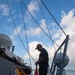 A naval sailor dressed in dark blue uniform and a cap is handling a large red and white flag on the deck of a military vessel. The ship appears to be underway or anchored near a coastline with green hills visible in the background. Multiple rigging lines, signal flags, and deck equipment are visible, consistent with a naval warship.
