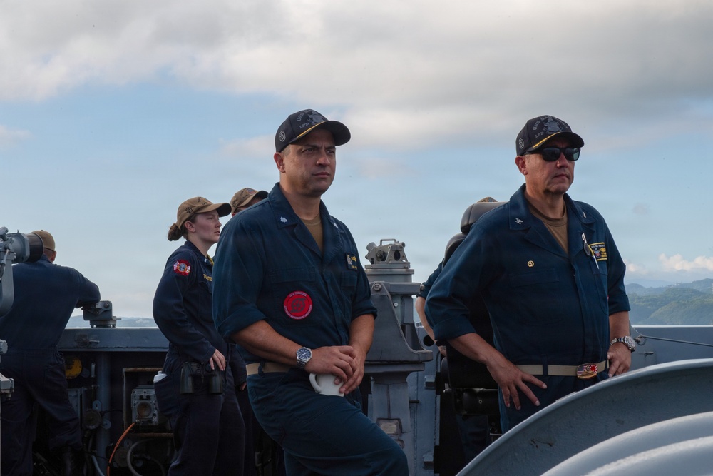 Several military personnel in navy blue uniforms are standing on the deck of a naval vessel. Two senior personnel are prominently positioned in the foreground, while additional crew members are visible in the background. The setting appears to be at sea with a coastline and partly cloudy sky visible in the background.