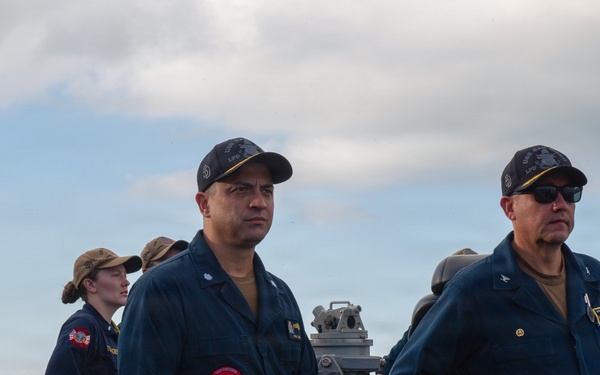 Several military personnel in navy blue uniforms are standing on the deck of a naval vessel. Two senior personnel are prominently positioned in the foreground, while additional crew members are visible in the background. The setting appears to be at sea with a coastline and partly cloudy sky visible in the background.