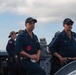 Several military personnel in navy blue uniforms are standing on the deck of a naval vessel. Two senior personnel are prominently positioned in the foreground, while additional crew members are visible in the background. The setting appears to be at sea with a coastline and partly cloudy sky visible in the background.