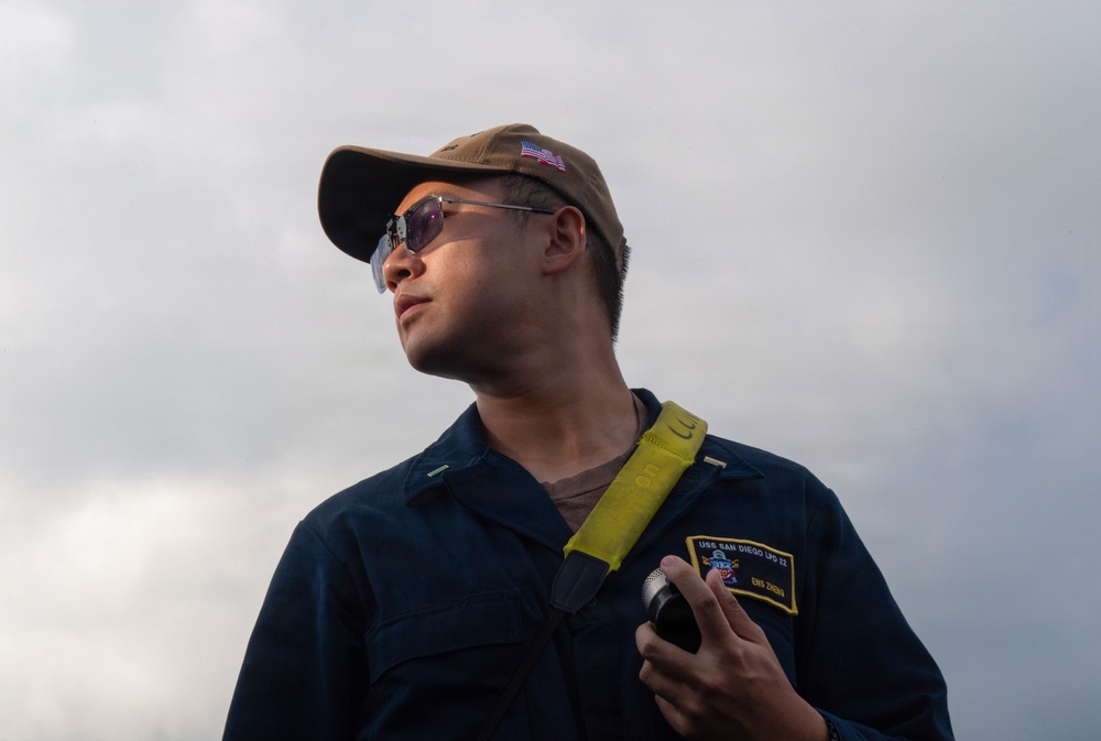 A U.S. Navy sailor wearing a dark blue uniform coverall with a name patch reading 'USS San Diego LPD 22' and rank 'ENS ZREBU' is photographed from a low angle against an overcast sky. He is wearing a tan baseball cap with a U.S. flag patch, tinted sunglasses, and has a yellow lanyard around his neck while holding a cylindrical object. The image appears to be an official or documentary military photograph.