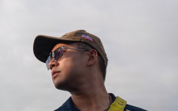 A U.S. Navy sailor wearing a dark blue uniform coverall with a name patch reading 'USS San Diego LPD 22' and rank 'ENS ZREBU' is photographed from a low angle against an overcast sky. He is wearing a tan baseball cap with a U.S. flag patch, tinted sunglasses, and has a yellow lanyard around his neck while holding a cylindrical object. The image appears to be an official or documentary military photograph.