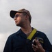 A U.S. Navy sailor wearing a dark blue uniform coverall with a name patch reading 'USS San Diego LPD 22' and rank 'ENS ZREBU' is photographed from a low angle against an overcast sky. He is wearing a tan baseball cap with a U.S. flag patch, tinted sunglasses, and has a yellow lanyard around his neck while holding a cylindrical object. The image appears to be an official or documentary military photograph.