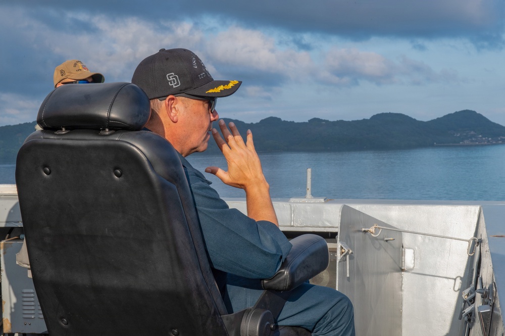 A person wearing a black San Diego Padres cap with gold insignia/scrambled eggs on the brim sits in a black leather command chair aboard what appears to be a naval vessel. They are holding a radio or communication device to their mouth while looking out toward a body of water with a mountainous island or coastline in the background. A second person wearing a tan/khaki cap is partially visible in the background.