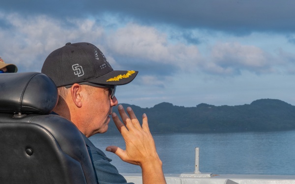 A person wearing a black San Diego Padres cap with gold insignia/scrambled eggs on the brim sits in a black leather command chair aboard what appears to be a naval vessel. They are holding a radio or communication device to their mouth while looking out toward a body of water with a mountainous island or coastline in the background. A second person wearing a tan/khaki cap is partially visible in the background.