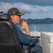 A person wearing a black San Diego Padres cap with gold insignia/scrambled eggs on the brim sits in a black leather command chair aboard what appears to be a naval vessel. They are holding a radio or communication device to their mouth while looking out toward a body of water with a mountainous island or coastline in the background. A second person wearing a tan/khaki cap is partially visible in the background.