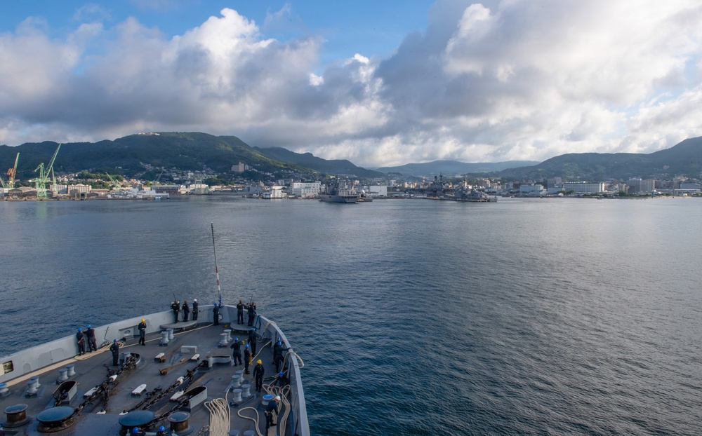 A naval vessel is approaching a harbor with a military installation visible along the shoreline, surrounded by green hills and a partially cloudy sky. Several crew members are stationed on the bow of the ship, likely performing arrival or mooring duties. Additional naval vessels are visible docked at the port in the background.