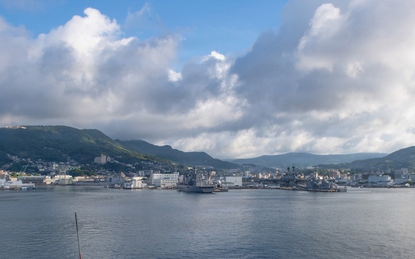 A naval vessel is approaching a harbor with a military installation visible along the shoreline, surrounded by green hills and a partially cloudy sky. Several crew members are stationed on the bow of the ship, likely performing arrival or mooring duties. Additional naval vessels are visible docked at the port in the background.