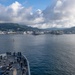 A naval vessel is approaching a harbor with a military installation visible along the shoreline, surrounded by green hills and a partially cloudy sky. Several crew members are stationed on the bow of the ship, likely performing arrival or mooring duties. Additional naval vessels are visible docked at the port in the background.