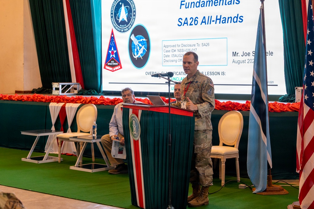 A military officer in camouflage uniform is presenting at a podium during what appears to be a U.S. Space Forces Europe Africa briefing event called 'SA26 All-Hands.' The presentation slide visible behind him references space fundamentals and is marked 'UNCLASSIFIED,' dated August 2022, with presenter identified as 'Mr. Joe S[omele/omething].' Two civilians are seated on stage behind the speaker, with U.S. and another nation's flags flanking the stage.