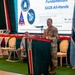A military officer in camouflage uniform is presenting at a podium during what appears to be a U.S. Space Forces Europe Africa briefing event called 'SA26 All-Hands.' The presentation slide visible behind him references space fundamentals and is marked 'UNCLASSIFIED,' dated August 2022, with presenter identified as 'Mr. Joe S[omele/omething].' Two civilians are seated on stage behind the speaker, with U.S. and another nation's flags flanking the stage.