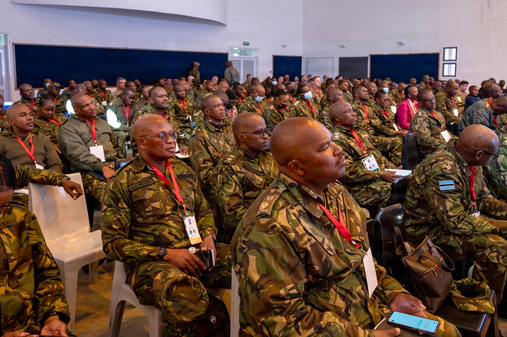 A large gathering of military personnel seated in rows inside a conference or briefing hall. The majority are wearing camouflage uniforms with red lanyards and identification badges, suggesting a formal military conference or seminar. The room is filled with dozens of attendees, indicating a significant multi-unit or multinational military event.