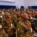 A large gathering of military personnel seated in rows inside a conference or briefing hall. The majority are wearing camouflage uniforms with red lanyards and identification badges, suggesting a formal military conference or seminar. The room is filled with dozens of attendees, indicating a significant multi-unit or multinational military event.