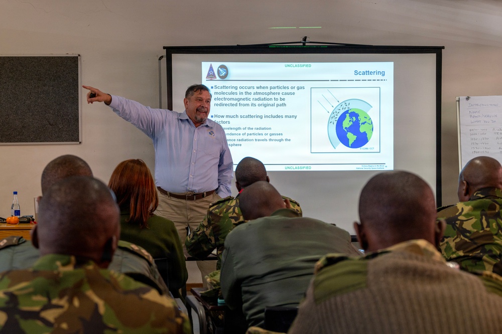 A civilian instructor is delivering a technical presentation on 'Scattering' of electromagnetic radiation to a group of military personnel in camouflage uniforms. The slide displayed is marked 'UNCLASSIFIED' and contains diagrams and bullet points explaining atmospheric scattering concepts. The setting appears to be a formal classroom or training facility.