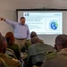 A civilian instructor is delivering a technical presentation on 'Scattering' of electromagnetic radiation to a group of military personnel in camouflage uniforms. The slide displayed is marked 'UNCLASSIFIED' and contains diagrams and bullet points explaining atmospheric scattering concepts. The setting appears to be a formal classroom or training facility.