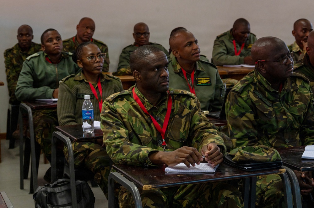A group of military personnel in camouflage uniforms are seated at desks in a classroom setting, appearing to attend a training or briefing session. All individuals are wearing red lanyards/badges, suggesting a formal organized event or course. The participants appear attentive, with some taking notes.