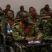 A group of military personnel in camouflage uniforms are seated at desks in a classroom setting, appearing to attend a training or briefing session. All individuals are wearing red lanyards/badges, suggesting a formal organized event or course. The participants appear attentive, with some taking notes.