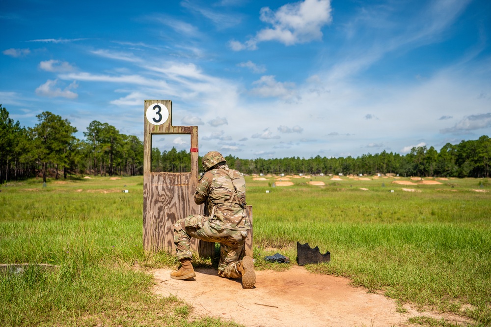 A soldier in full camouflage uniform and combat gear is kneeling at a numbered shooting station (position 3) on a military firing range. The shooter is in a kneeling position behind a wooden barricade, aiming downrange toward distant targets visible across an open grassy field. The setting appears to be a well-maintained outdoor military qualification or competition range with multiple target positions visible in the background.