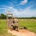 A soldier in full camouflage uniform and combat gear is kneeling at a numbered shooting station (position 3) on a military firing range. The shooter is in a kneeling position behind a wooden barricade, aiming downrange toward distant targets visible across an open grassy field. The setting appears to be a well-maintained outdoor military qualification or competition range with multiple target positions visible in the background.