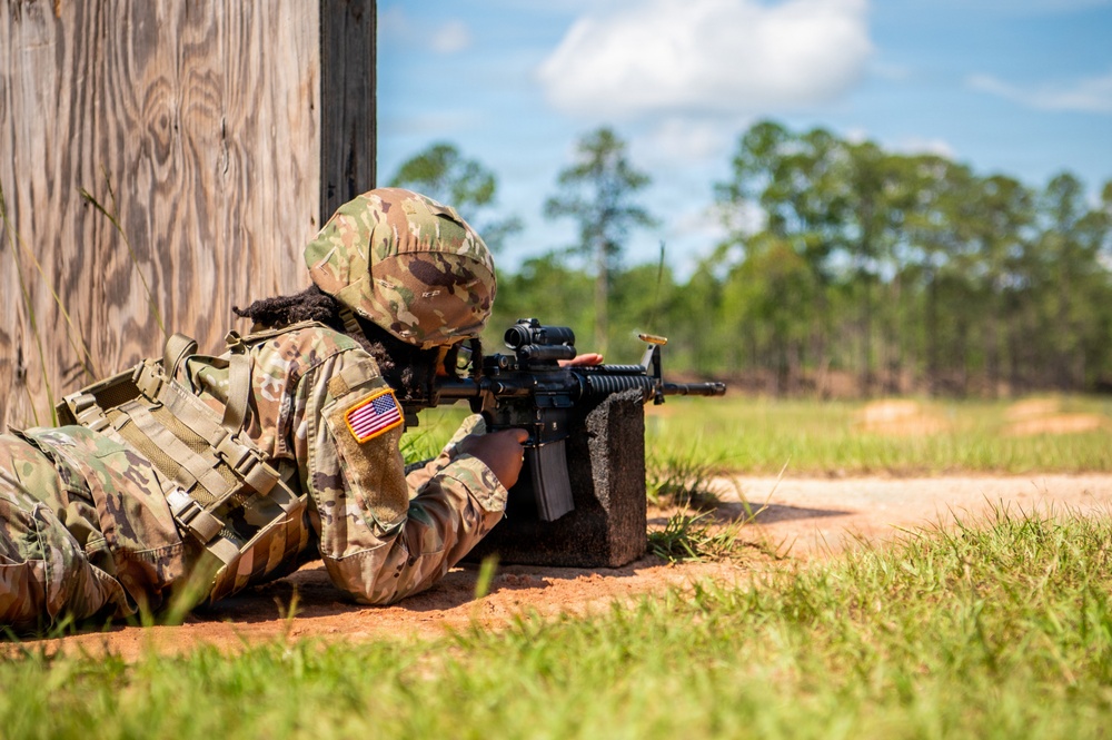 A U.S. Army soldier in full combat gear is in a prone firing position, aiming a rifle around the corner of a wooden structure. The soldier is wearing Operational Camouflage Pattern (OCP) uniform with a visible American flag patch on the left sleeve. The setting appears to be a military training range in a warm, wooded environment.