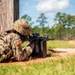 A U.S. Army soldier in full combat gear is in a prone firing position, aiming a rifle around the corner of a wooden structure. The soldier is wearing Operational Camouflage Pattern (OCP) uniform with a visible American flag patch on the left sleeve. The setting appears to be a military training range in a warm, wooded environment.