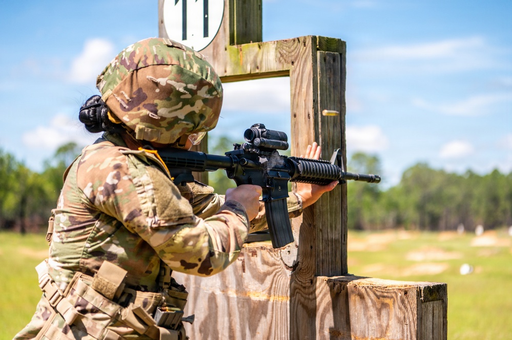 A soldier in full OCP (Operational Camouflage Pattern) uniform is actively firing an M4/M16-style carbine rifle from behind a wooden barricade on an outdoor shooting range. A spent brass casing is visibly ejecting from the weapon, indicating the rifle has just been fired. The setting appears to be a military marksmanship training or qualification range during daytime.