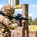 A soldier in full OCP (Operational Camouflage Pattern) uniform is actively firing an M4/M16-style carbine rifle from behind a wooden barricade on an outdoor shooting range. A spent brass casing is visibly ejecting from the weapon, indicating the rifle has just been fired. The setting appears to be a military marksmanship training or qualification range during daytime.