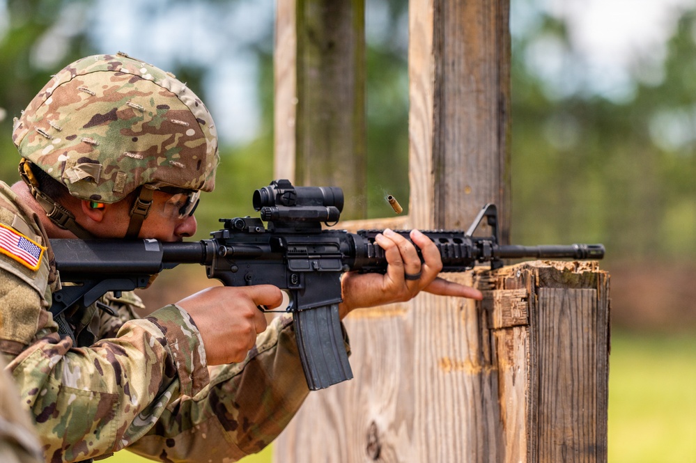 A U.S. Army soldier in Operational Camouflage Pattern (OCP) uniform is actively firing an M4 carbine rifle from a covered position behind a wooden barrier. A spent brass casing is visibly ejecting from the weapon's ejection port, indicating the weapon has just been fired. The setting appears to be an outdoor military training range.