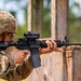 A U.S. Army soldier in Operational Camouflage Pattern (OCP) uniform is actively firing an M4 carbine rifle from a covered position behind a wooden barrier. A spent brass casing is visibly ejecting from the weapon's ejection port, indicating the weapon has just been fired. The setting appears to be an outdoor military training range.
