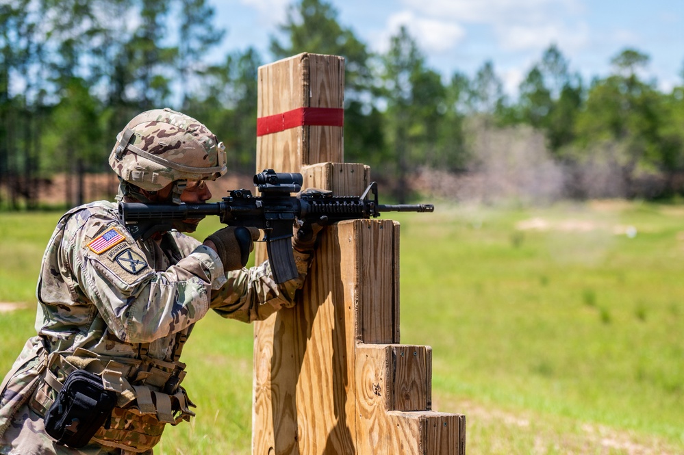 A U.S. Army soldier in full combat gear is firing an M4 carbine rifle from behind a wooden barricade on a training range. The soldier is wearing Operational Camouflage Pattern (OCP) uniform with visible U.S. flag and 10th Mountain Division patches. Dust or smoke is visible downrange, consistent with an active firing exercise.