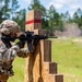 A U.S. Army soldier in full combat gear is firing an M4 carbine rifle from behind a wooden barricade on a training range. The soldier is wearing Operational Camouflage Pattern (OCP) uniform with visible U.S. flag and 10th Mountain Division patches. Dust or smoke is visible downrange, consistent with an active firing exercise.