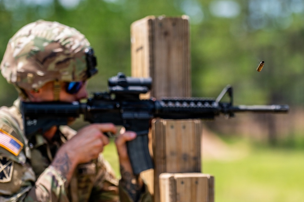 A U.S. Army soldier in MultiCam (OCP) uniform is firing an M4 carbine rifle at a shooting range, using a wooden barrier as cover. A brass cartridge casing is visibly ejecting from the weapon, captured mid-air by the camera. The soldier is wearing a combat helmet, blue-tinted eye protection, and has a U.S. flag patch and what appears to be a 1st Cavalry Division patch on the uniform.
