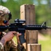 A U.S. Army soldier in MultiCam (OCP) uniform is firing an M4 carbine rifle at a shooting range, using a wooden barrier as cover. A brass cartridge casing is visibly ejecting from the weapon, captured mid-air by the camera. The soldier is wearing a combat helmet, blue-tinted eye protection, and has a U.S. flag patch and what appears to be a 1st Cavalry Division patch on the uniform.