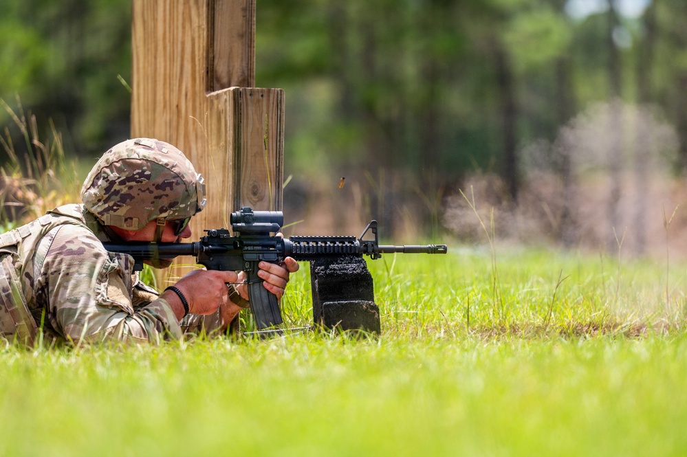 A soldier in full OCP (Operational Camouflage Pattern) uniform is firing an M4 carbine rifle from a prone position on a grass range. A spent brass casing is visibly ejecting from the weapon, indicating active firing. The soldier is using a wooden barrier as cover during what appears to be a military marksmanship training exercise.