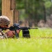 A soldier in full OCP (Operational Camouflage Pattern) uniform is firing an M4 carbine rifle from a prone position on a grass range. A spent brass casing is visibly ejecting from the weapon, indicating active firing. The soldier is using a wooden barrier as cover during what appears to be a military marksmanship training exercise.