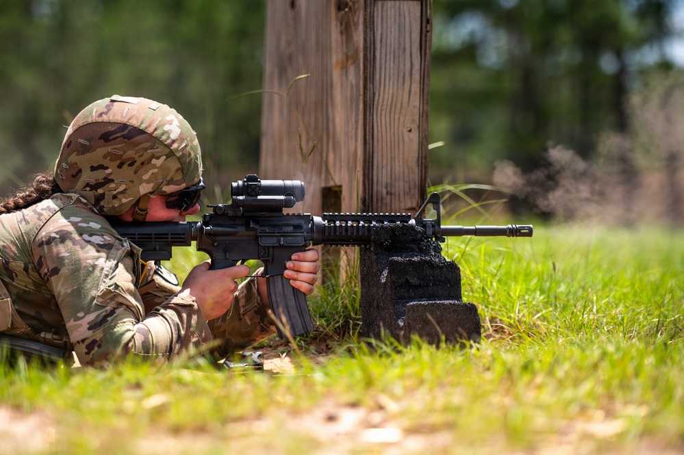 A soldier in full OCP (Operational Camouflage Pattern) uniform is in a prone firing position on a grass range, aiming an M4/M16-style carbine with an optic and rail-mounted accessories. The shooter is using a concrete block as a rest or barricade support near a wooden post. The setting appears to be an outdoor military training range.