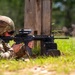 A soldier in full OCP (Operational Camouflage Pattern) uniform is in a prone firing position on a grass range, aiming an M4/M16-style carbine with an optic and rail-mounted accessories. The shooter is using a concrete block as a rest or barricade support near a wooden post. The setting appears to be an outdoor military training range.