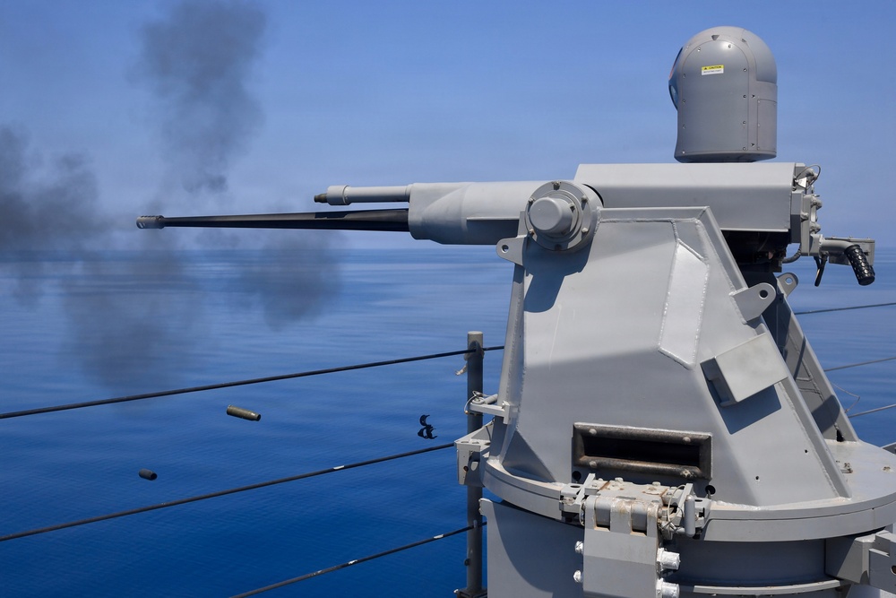 A naval deck-mounted automatic cannon system is actively firing over open blue water. Spent brass casings are visibly ejecting from the weapon, and a plume of dark smoke is visible in the background, likely from a target or previous rounds. The system appears to be a remotely operated naval gun mount installed on the deck of a warship.