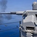 A naval deck-mounted automatic cannon system is actively firing over open blue water. Spent brass casings are visibly ejecting from the weapon, and a plume of dark smoke is visible in the background, likely from a target or previous rounds. The system appears to be a remotely operated naval gun mount installed on the deck of a warship.