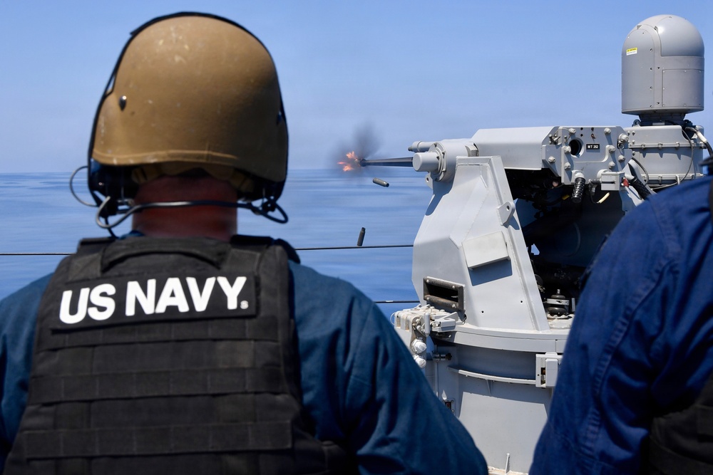 A US Navy sailor wearing a tactical vest and ballistic helmet observes a deck-mounted naval gun system firing during what appears to be a live-fire exercise at sea. A muzzle flash and ejected brass casing are visible, indicating the weapon is actively firing. The setting is a naval vessel on open, calm blue water under clear skies.