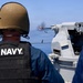 A US Navy sailor wearing a tactical vest and ballistic helmet observes a deck-mounted naval gun system firing during what appears to be a live-fire exercise at sea. A muzzle flash and ejected brass casing are visible, indicating the weapon is actively firing. The setting is a naval vessel on open, calm blue water under clear skies.