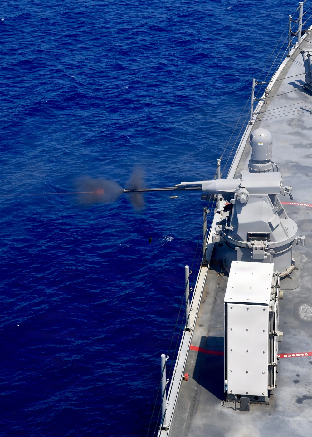 An aerial photograph captures a naval warship firing a deck-mounted gun over open ocean. The weapon system is actively discharging, with visible muzzle flash, smoke, and what appears to be a spent casing ejecting near the water. The ship's gray hull, deck equipment, and safety markings are clearly visible from the elevated vantage point.