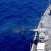 An aerial photograph captures a naval warship firing a deck-mounted gun over open ocean. The weapon system is actively discharging, with visible muzzle flash, smoke, and what appears to be a spent casing ejecting near the water. The ship's gray hull, deck equipment, and safety markings are clearly visible from the elevated vantage point.