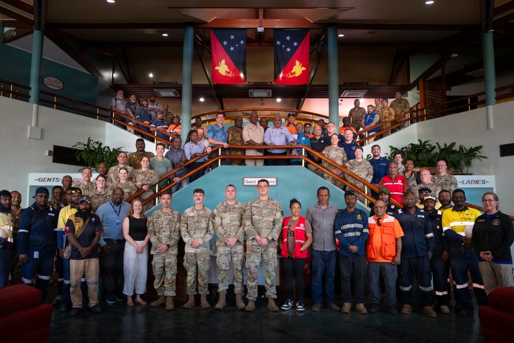 A large group photo taken inside a building featuring military personnel in camouflage uniforms, civilian workers in high-visibility safety gear, and officials posing across multiple levels of a staircase. The Papua New Guinea national flag is prominently displayed in the background, suggesting this is an official or governmental venue in PNG. The gathering appears to represent a multinational or inter-agency collaboration involving military, humanitarian, and civilian organizations.