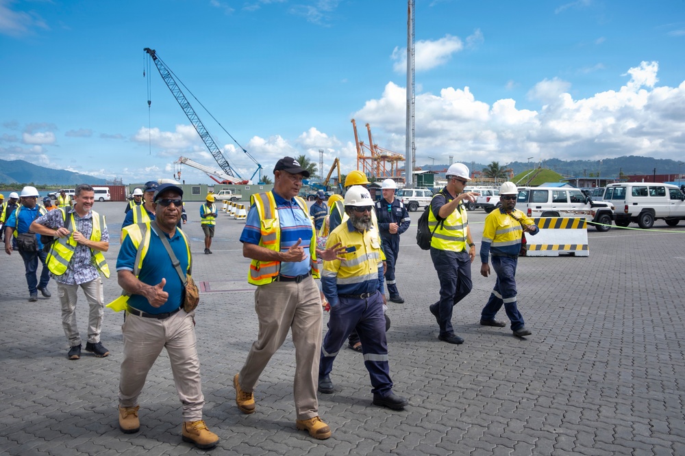 A group of workers and officials are conducting a site walkthrough or inspection at what appears to be an industrial port facility. Multiple individuals are wearing high-visibility safety vests and hard hats, suggesting a formal safety-compliant site visit. The background reveals port infrastructure including cranes, shipping containers, and heavy equipment consistent with a working cargo terminal.