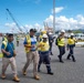 A group of workers and officials are conducting a site walkthrough or inspection at what appears to be an industrial port facility. Multiple individuals are wearing high-visibility safety vests and hard hats, suggesting a formal safety-compliant site visit. The background reveals port infrastructure including cranes, shipping containers, and heavy equipment consistent with a working cargo terminal.