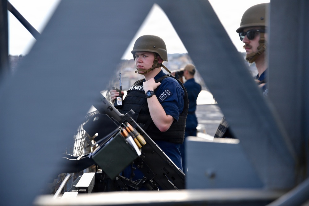 Two U.S. Navy personnel are stationed on what appears to be the deck of a naval vessel, photographed through a metal lattice structure. The foreground sailor is wearing a combat helmet, body armor vest, and a blue U.S. Navy shirt while holding a radio, positioned behind a mounted machine gun with visible ammunition. A second sailor wearing sunglasses and a tan helmet is visible in the background.