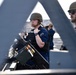 Two U.S. Navy personnel are stationed on what appears to be the deck of a naval vessel, photographed through a metal lattice structure. The foreground sailor is wearing a combat helmet, body armor vest, and a blue U.S. Navy shirt while holding a radio, positioned behind a mounted machine gun with visible ammunition. A second sailor wearing sunglasses and a tan helmet is visible in the background.