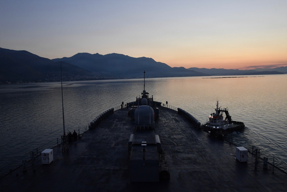 The image shows the bow of a large naval or commercial vessel photographed from an elevated stern perspective during twilight or early dawn. A tugboat with illuminated lights is positioned alongside the starboard side of the main vessel, likely assisting with maneuvering. A mountainous coastline with a small settlement is visible in the background across calm waters.